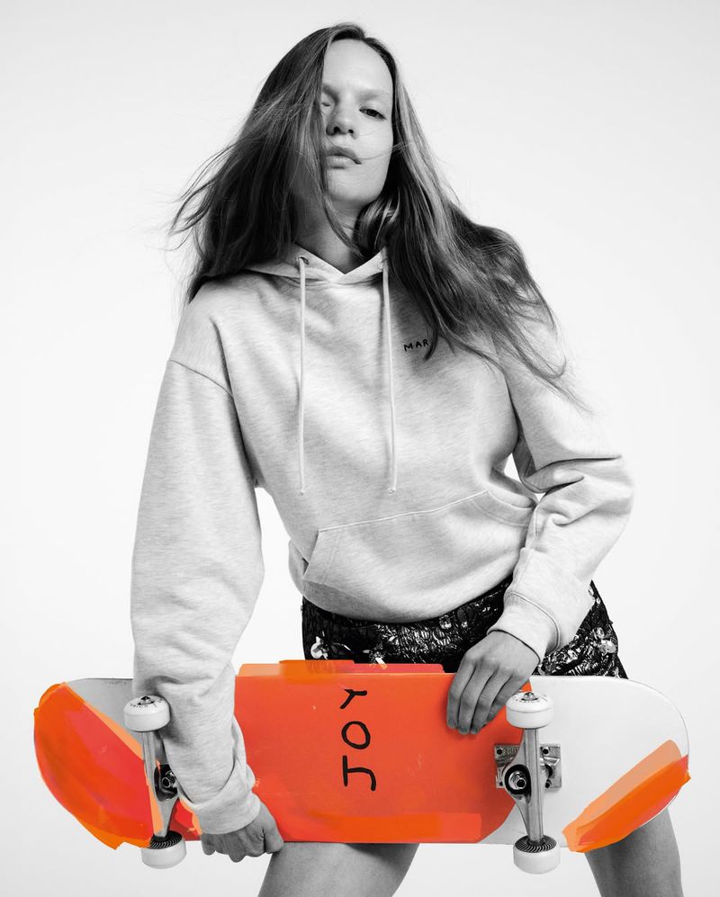 A young adult woman poses in a grey hoodie and black shorts, holding a bright orange and white skateboard against a white backdrop.