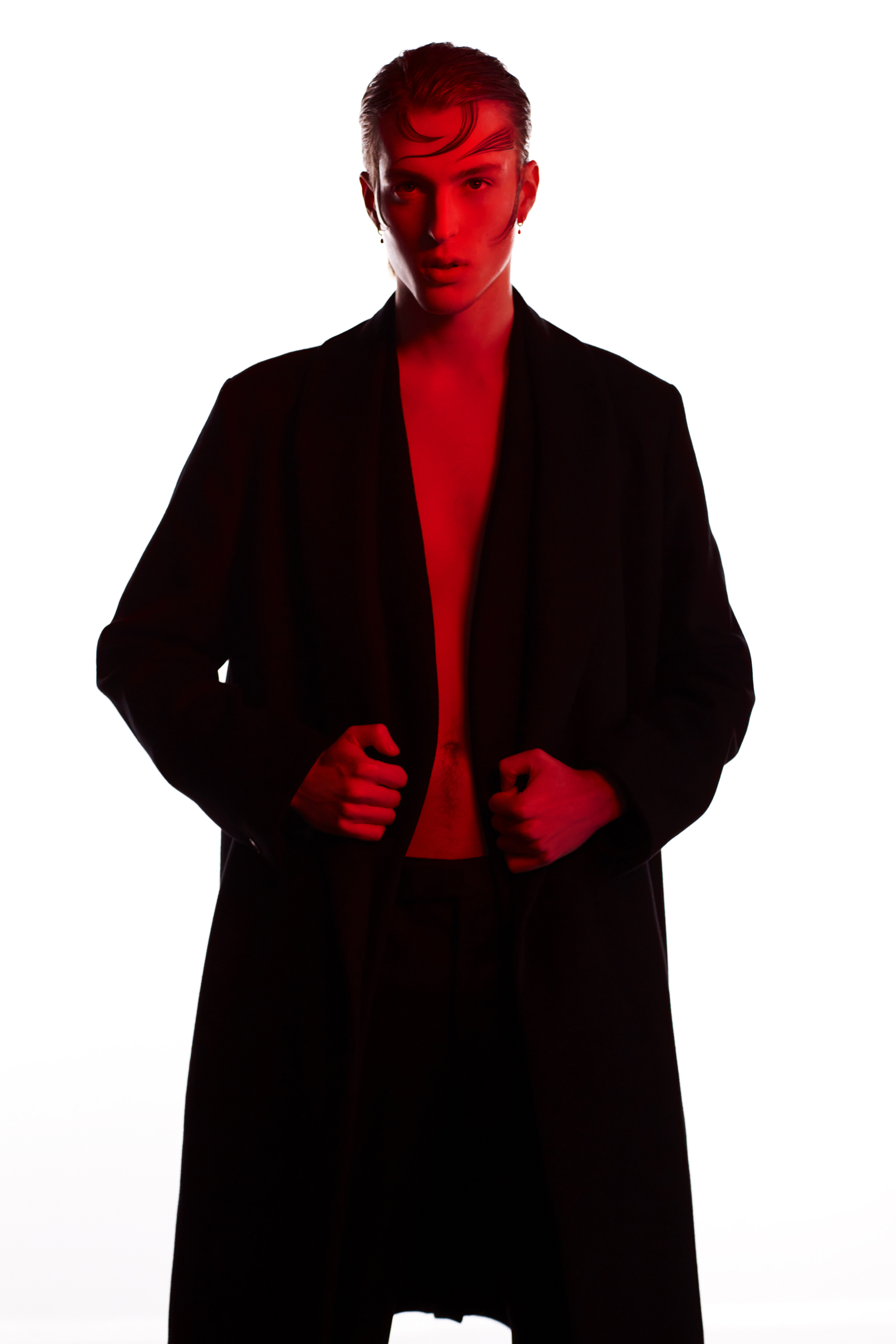 A stylish young adult stands indoors wearing a dark coat with red lighting effects against a bright white backdrop, showcasing a fashionable look.
