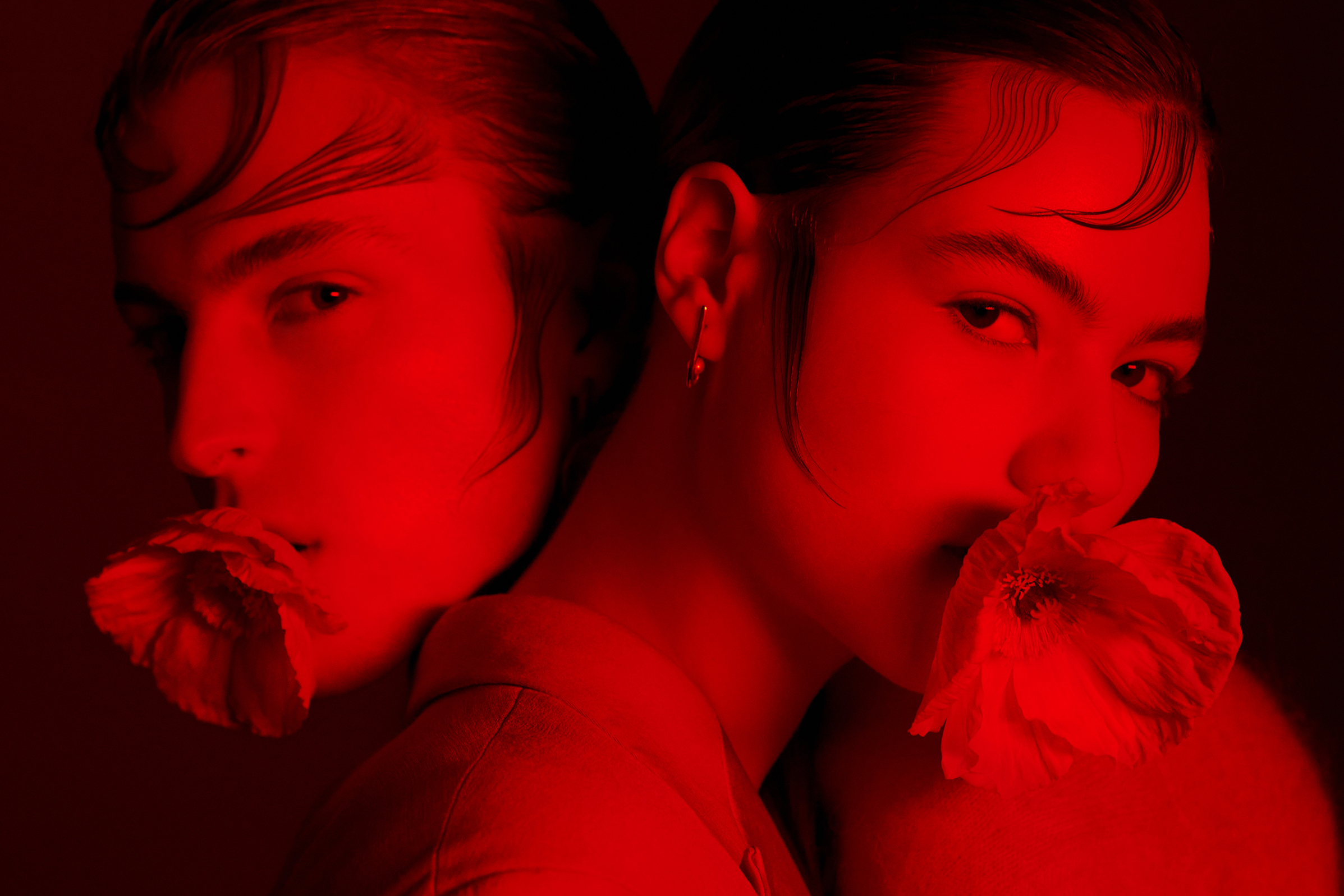 Portrait of two young adults in a close-up shot, bathed in saturated red lighting, each holding a flower to their mouths in a studio.
