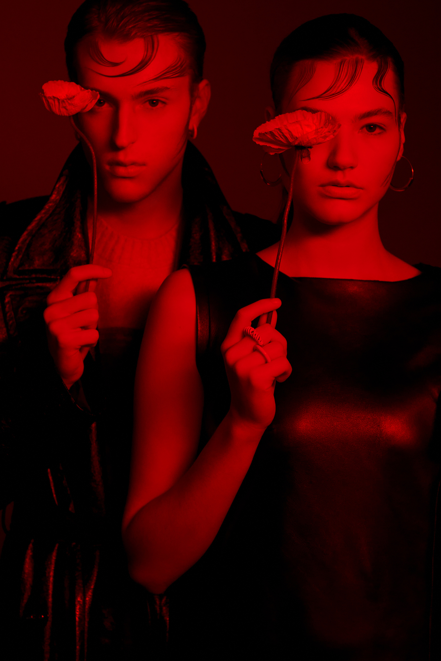 A striking portrait of a couple with sleek hair, bathed in a dark red glow, each holding a delicate flower in front of their face.