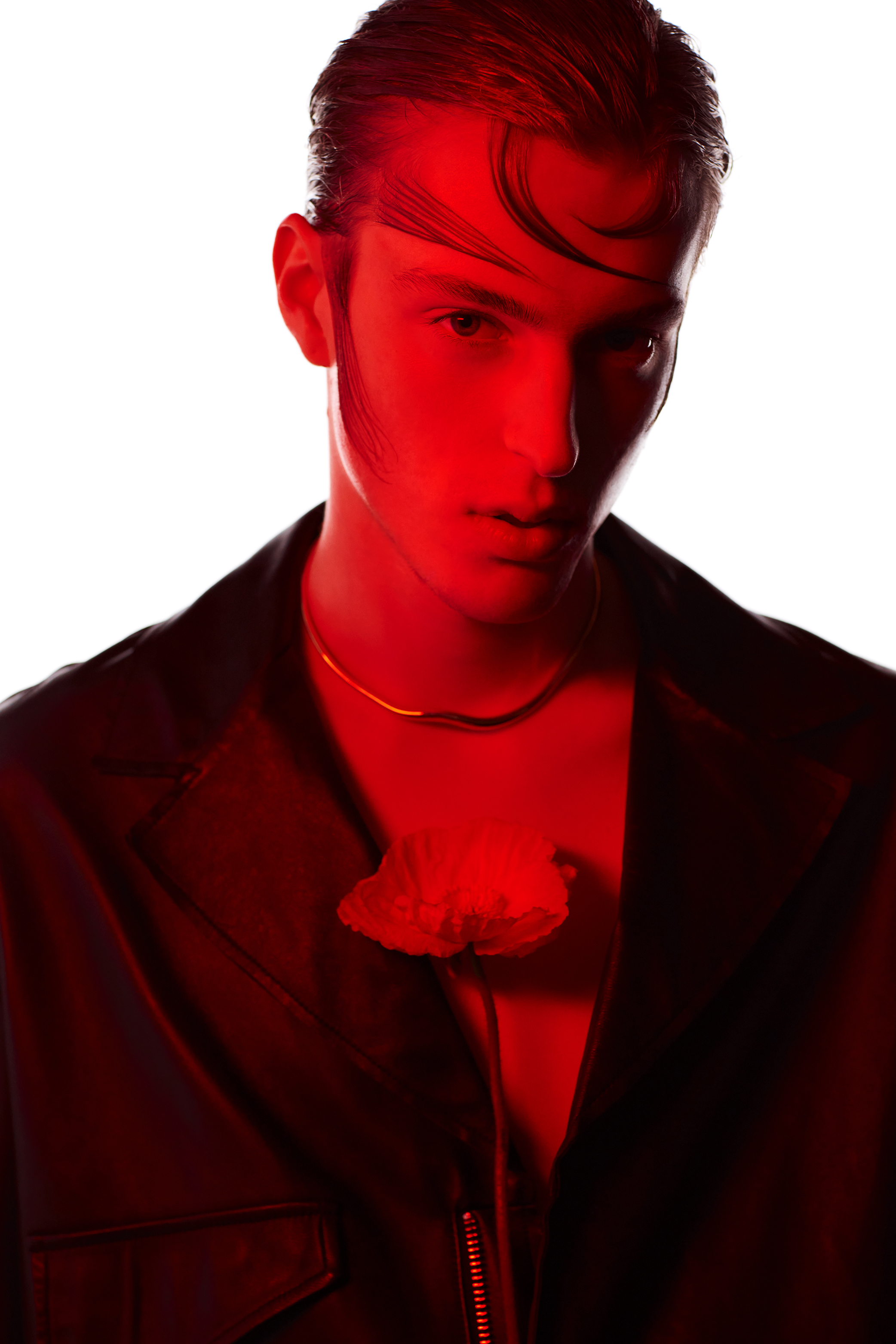 Close-up of a young adult male in a black jacket, illuminated by red light against a bright white background. He wears a necklace.
