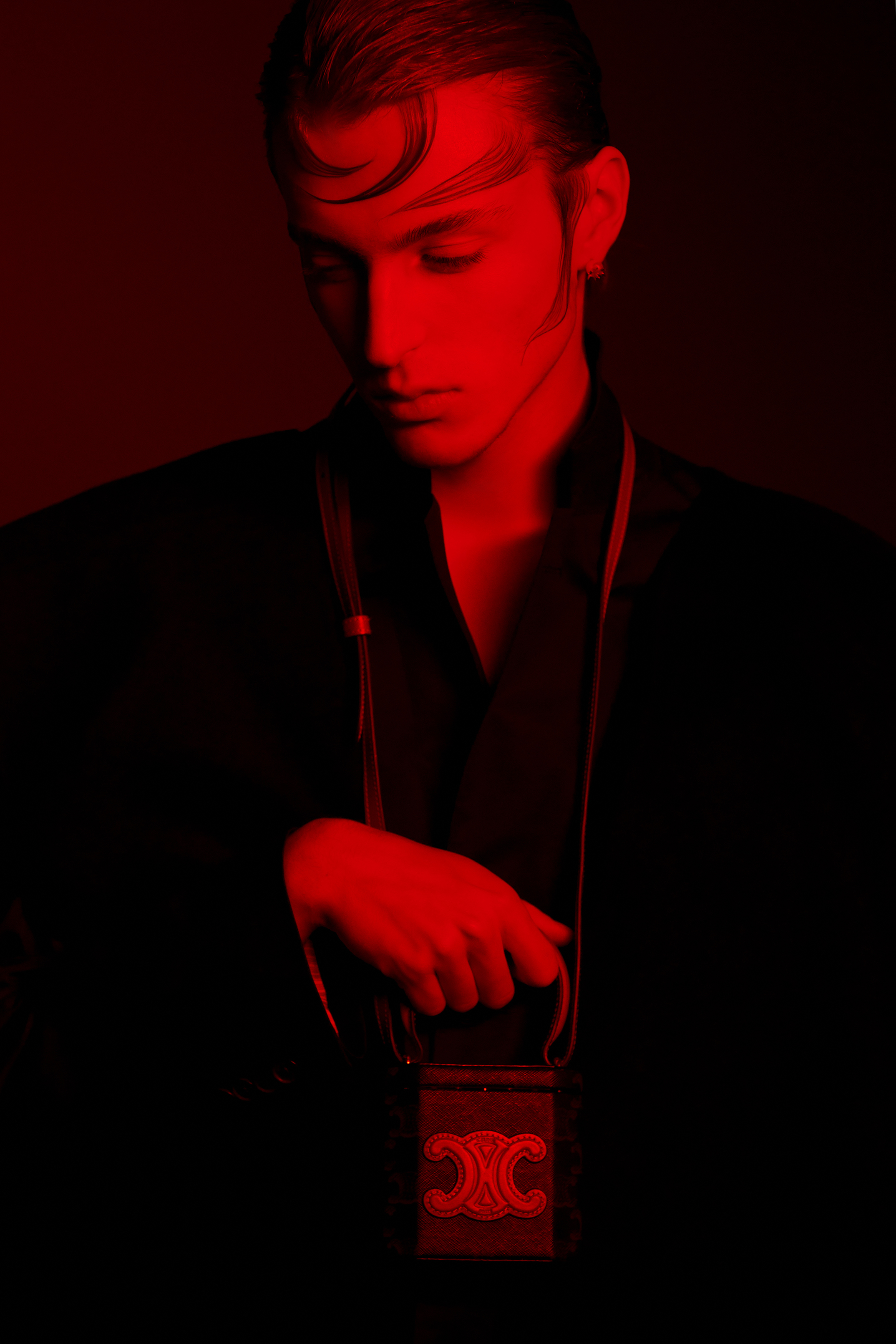 Portrait of a stylish adult male in a black jacket, holding a black Celine purse with gold accents under a red studio light.