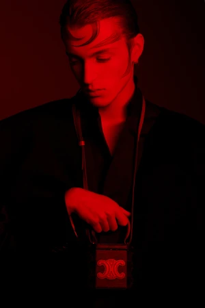 Portrait of a stylish adult male in a black jacket, holding a black Celine purse with gold accents under a red studio light.
