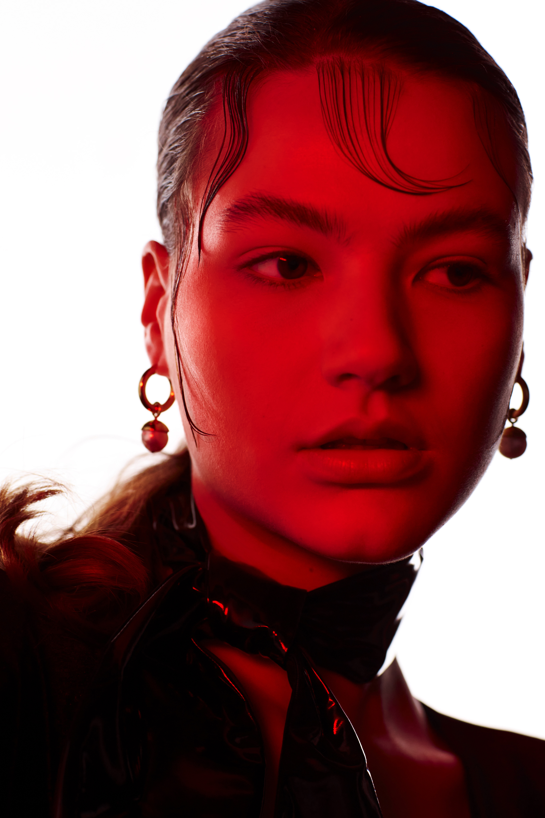 Close-up studio portrait of an Asian woman with wet-look hair, gold hoop earrings, and a dark top against a bright white background.