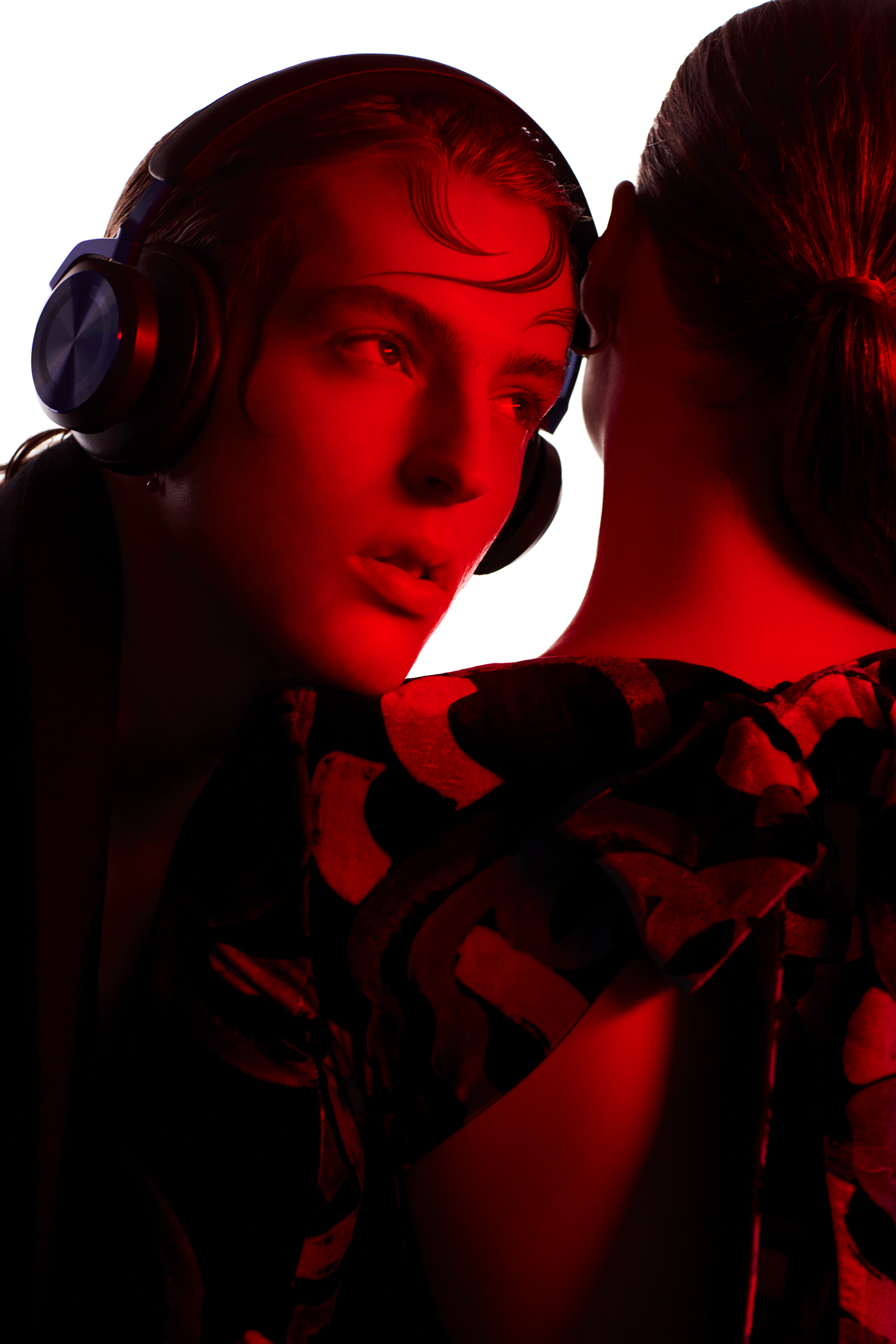 Two models in a close-up studio shot. One wears headphones, and both are illuminated with red lighting against a white background.
