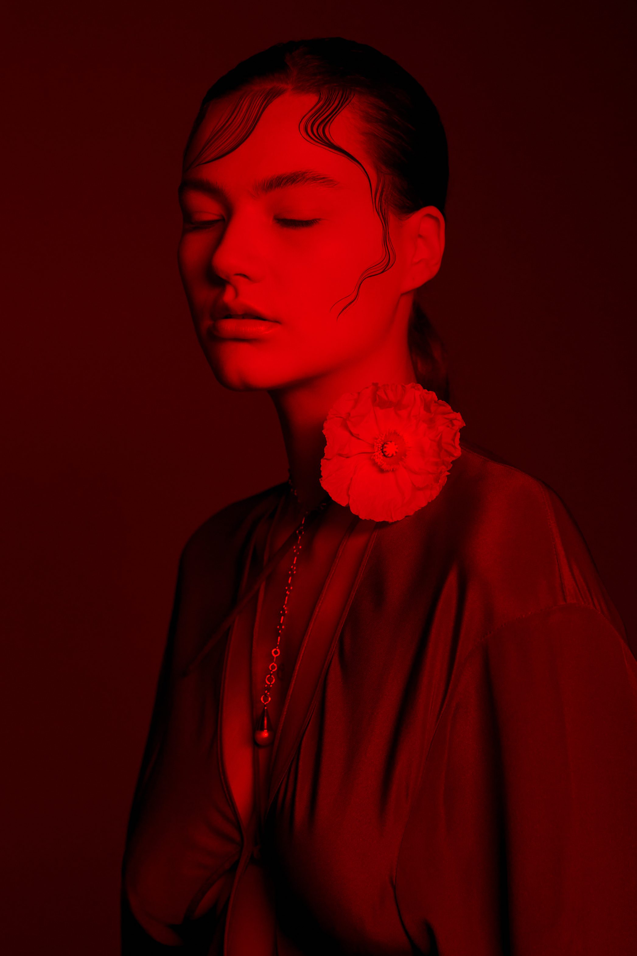 Portrait of a fair-skinned young adult woman with closed eyes in vibrant red lighting, wearing a flowing blouse and flower on the shoulder.