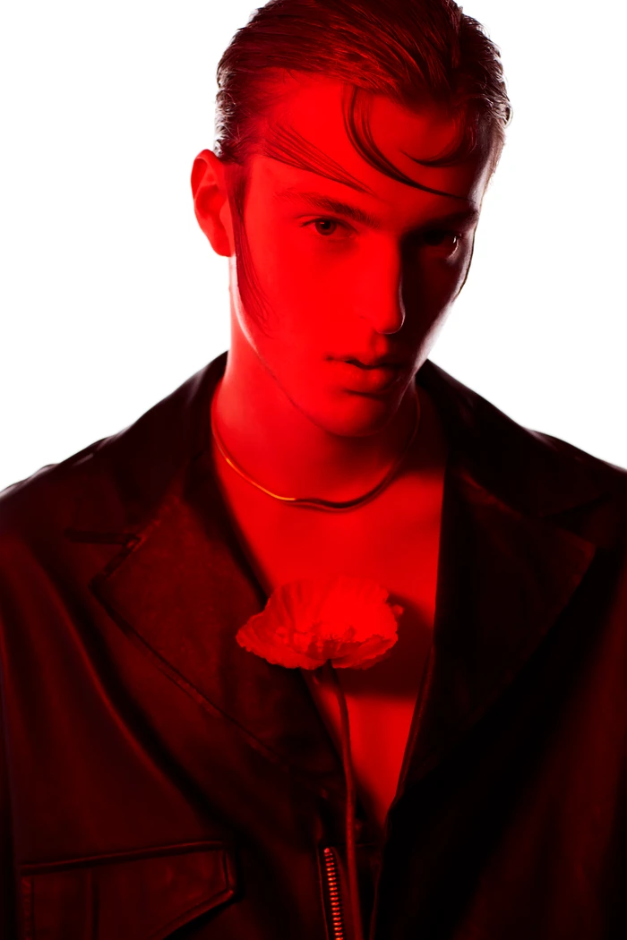 Close-up portrait of a male model bathed in red light, wearing a dark jacket with a single flower tucked into the collar.