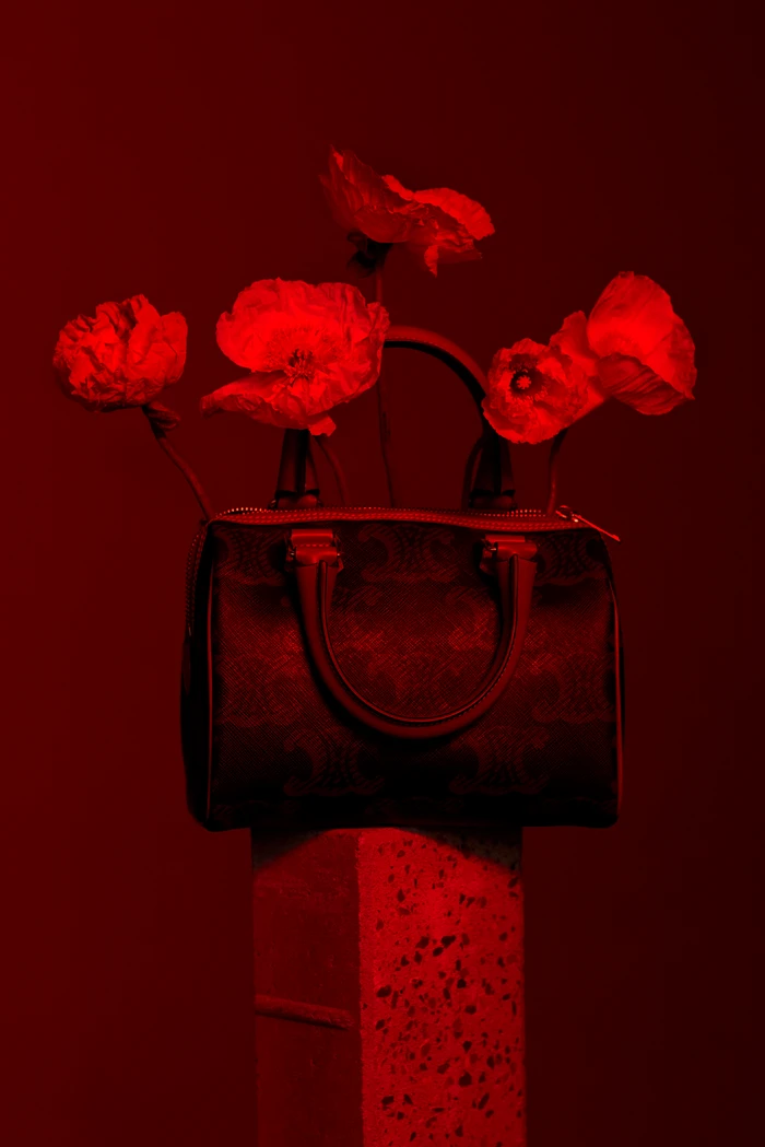 Dark handbag resting on a pedestal with red poppies emerging from it under deep red lighting