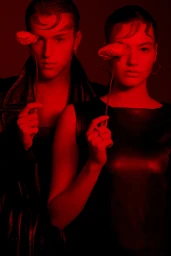Two models bathed in deep red light holding poppies over one eye for Base cast by Milos Mlynarik.