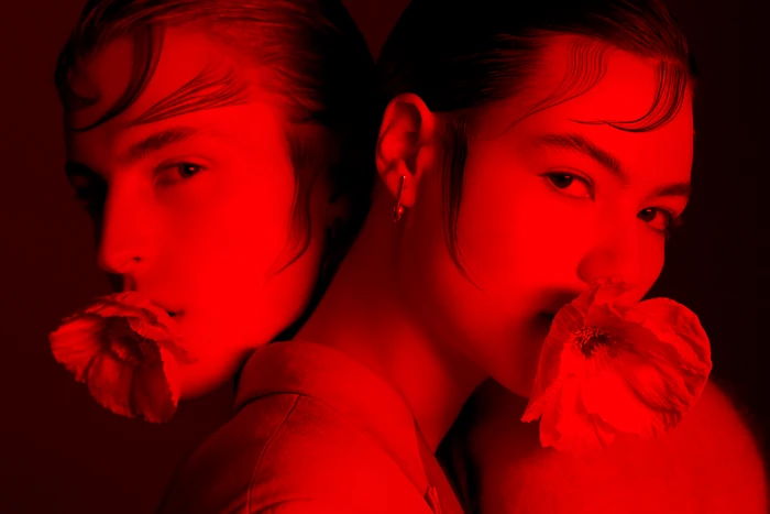 Two models posed closely together against a dark red backdrop, each holding a poppy flower in their mouth under vivid red lighting