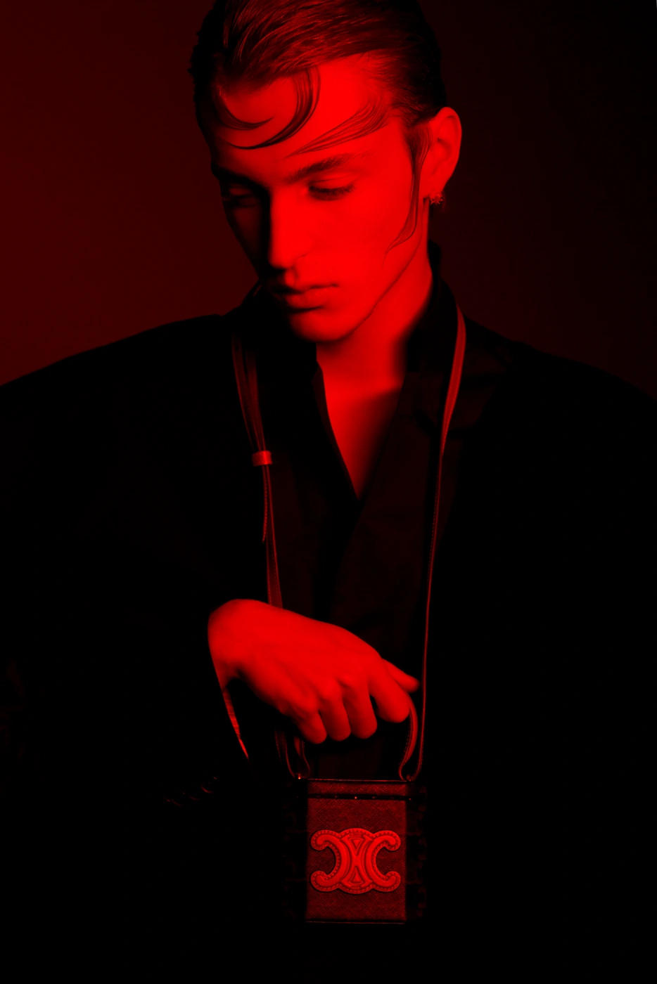 Portrait of a man in dark clothing bathed in intense red light, holding a small structured handbag with a prominent clasp.