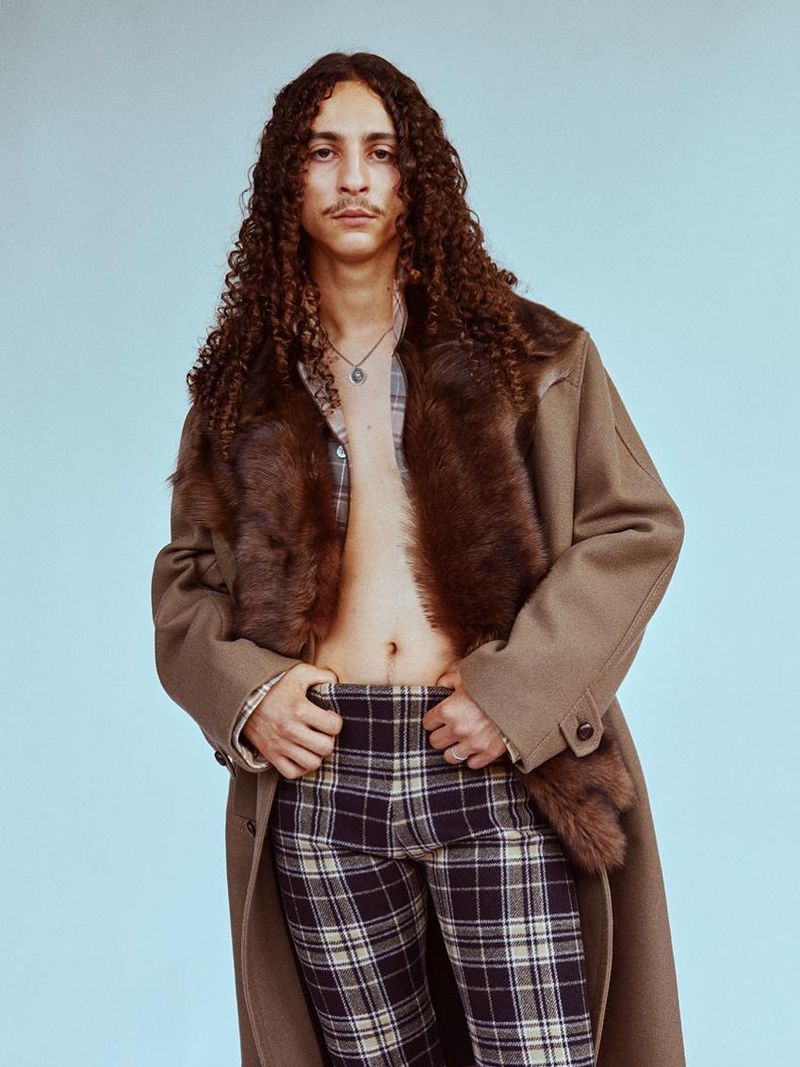 Portrait of a person with long, curly brown hair, wearing a tan coat with a fur collar and plaid pants against a light blue background.