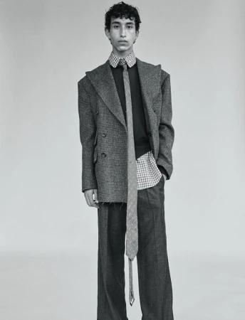 Male fashion model poses in an oversized grey tweed suit with a long tie. Black and white studio shot against a neutral backdrop.