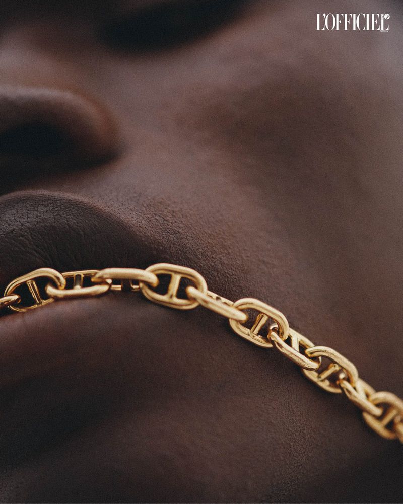 Close up shot of dark skin model wearing gold chain link necklace against a neutral background for fashion editorial with L'Officiel logo.
