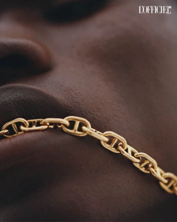 Close up shot of dark skin model wearing gold chain link necklace against a neutral background for fashion editorial with L'Officiel logo.