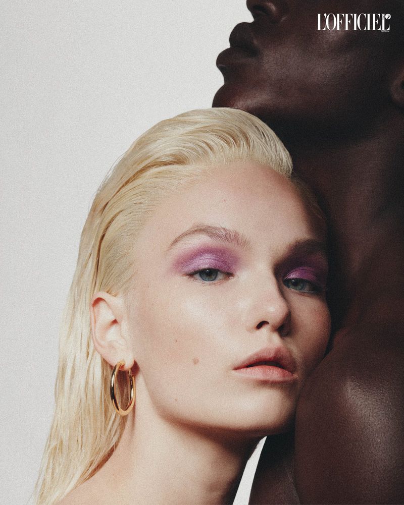Close-up of a fair-skinned woman with platinum blonde hair and a darker-skinned man partially visible in a beauty-style editorial shot.
