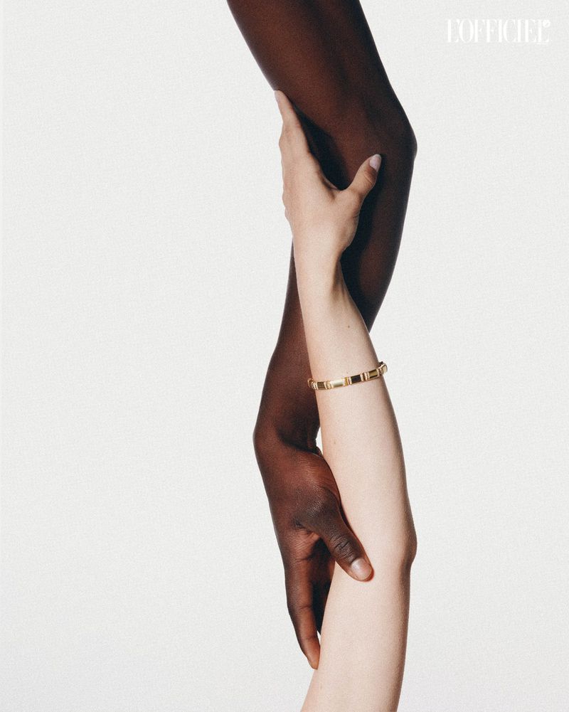 Close-up of a dark-skinned arm and a light-skinned arm touching, showcasing a gold bracelet against a bright, minimalist background.