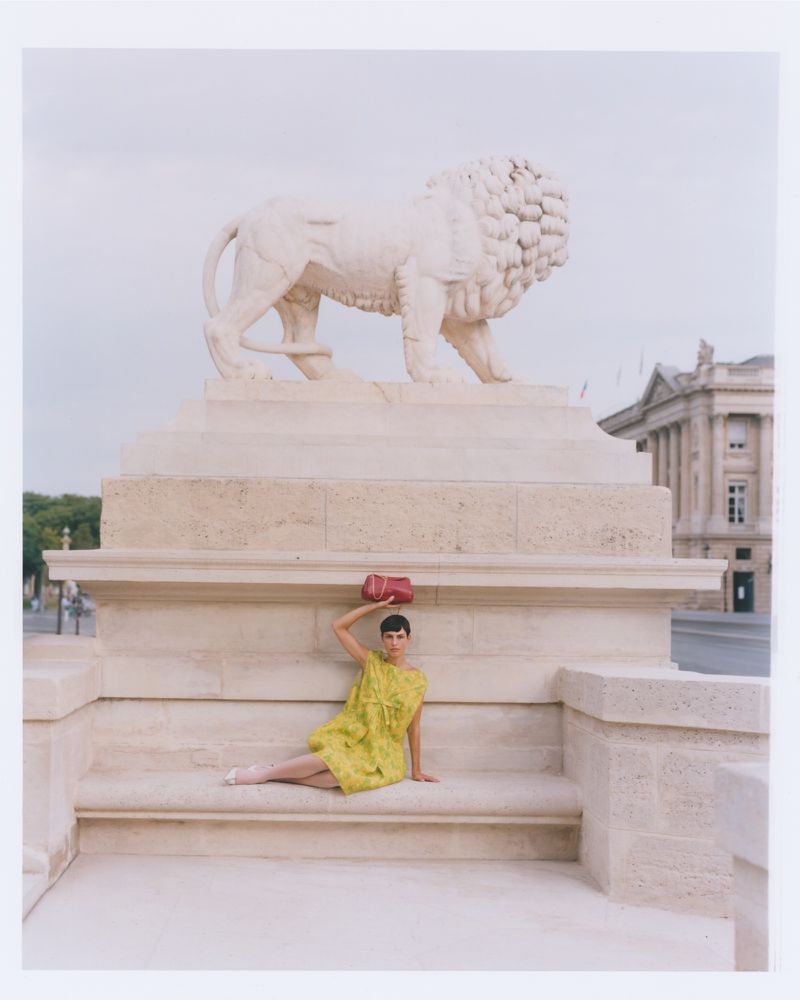 Woman in a yellow dress poses with a red handbag on her head in front of a stone lion statue and Parisian architecture.