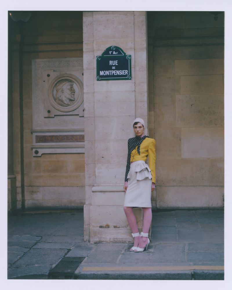 Fashion shoot of a model in a yellow and black jacket, cream skirt, and pink tights standing on a street with a building in the background.
