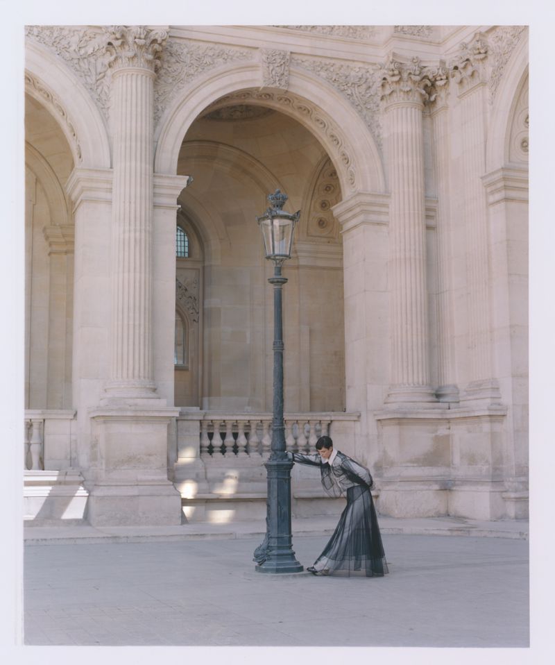 Woman in gray leans against a black lamppost in a tan stone courtyard with arched openings and fluted columns on a sunny day.