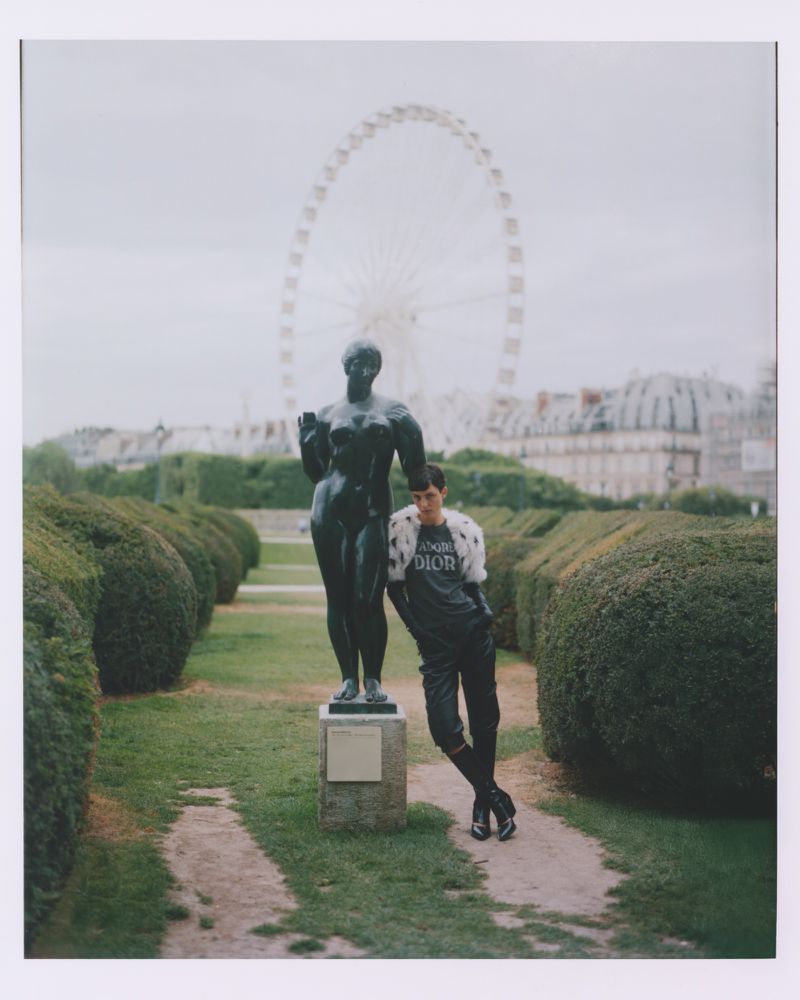 Adult male poses next to a bronze statue in a park with trimmed hedges and a distant Ferris wheel. The scene is captured in daylight.