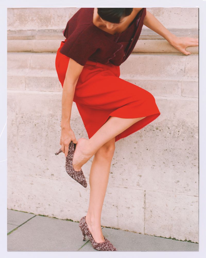 Adult woman in red skirt adjusts a patterned pump in a polaroid-style shot against a light neutral wall with architectural details.