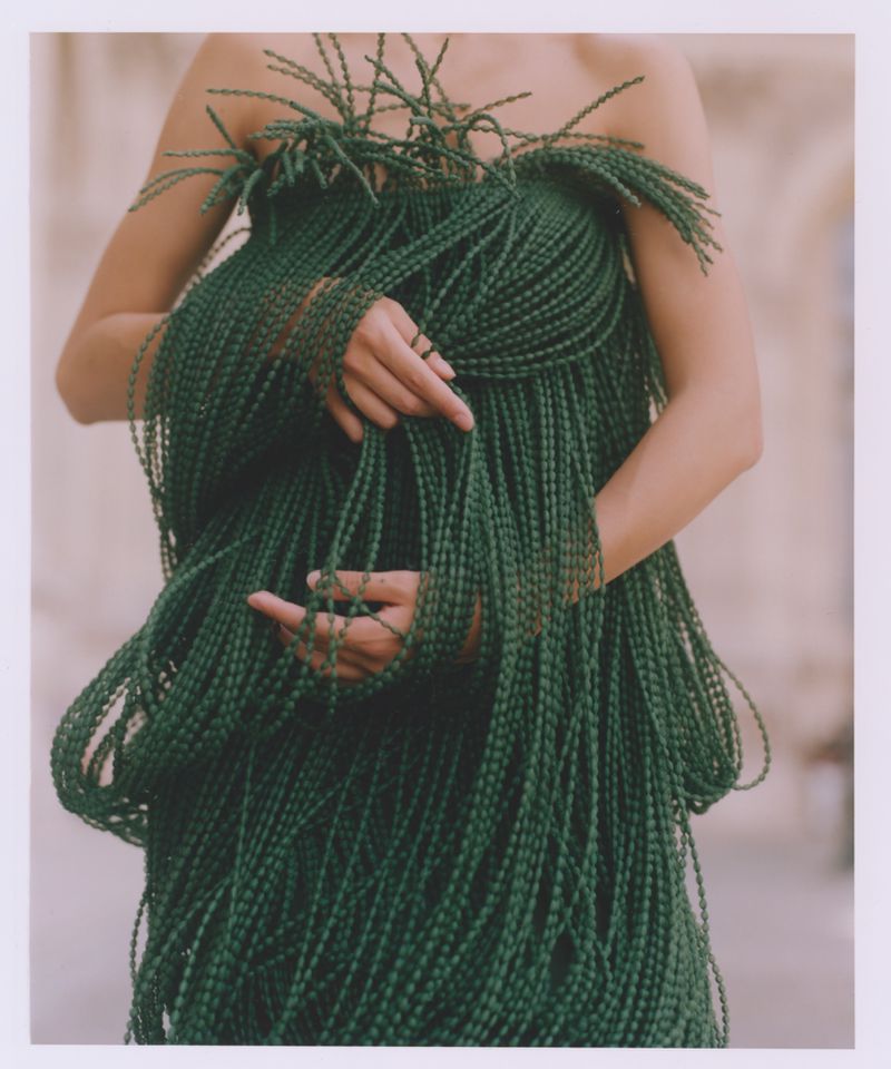 Woman in green beaded dress, standing in front of blurred-out building, facing forward with her hands in the center of the frame.