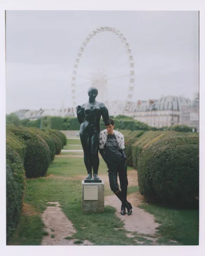 Model Mary Alexandridi leaning against a bronze statue in a formal park with manicured hedges and a distant Ferris wheel.