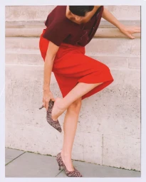 Model Mary Alexandridi in a red skirt and burgundy top adjusting her patterned pointed heel against an outdoor stone wall.
