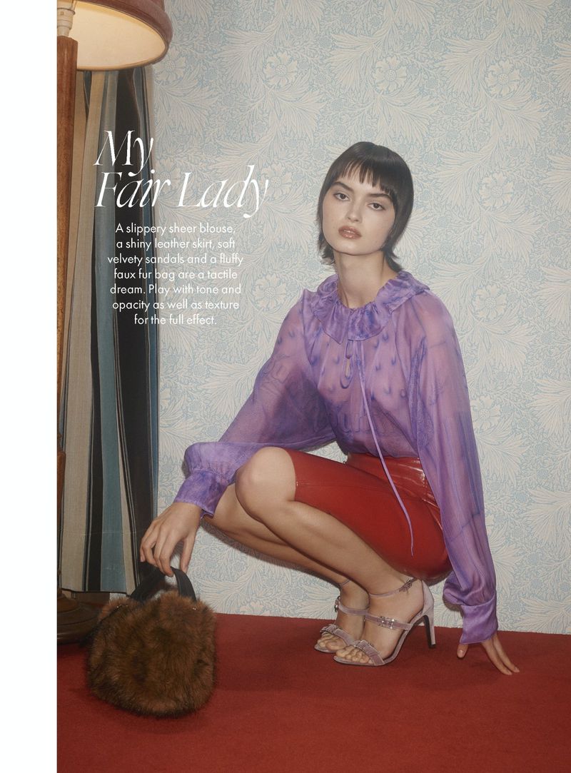 A young woman poses in a sheer blouse and red skirt, kneeling on a carpet in a fashion editorial shot. She holds a furry bag.