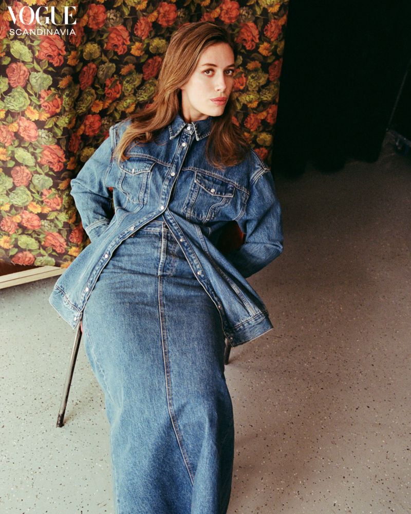A woman in a denim outfit poses on a chair against a floral backdrop, showcasing fashion and editorial style with natural lighting.