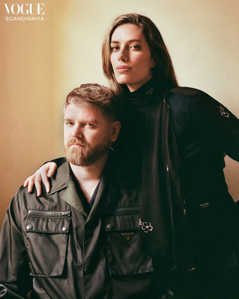 Editorial portrait of a male and female wearing Prada jackets indoors with soft lighting, posing together in front of a light-yellow background.