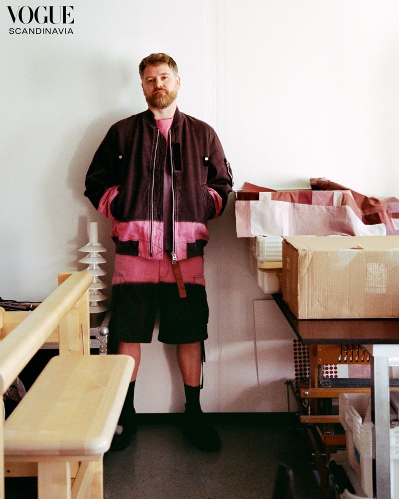 Man with a beard standing in a workshop wearing a two-tone brown and pink bomber jacket and matching shorts.