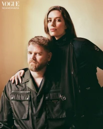 A bearded man seated beside a woman resting her hand on his shoulder, both wearing black outerwear against a warm wall.