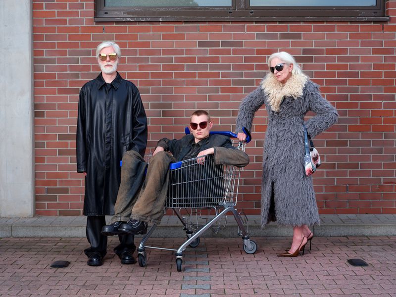 Three people pose in front of a brick wall: an older man in a black trench coat, a young adult in a shopping cart, and an older woman in a fur coat.