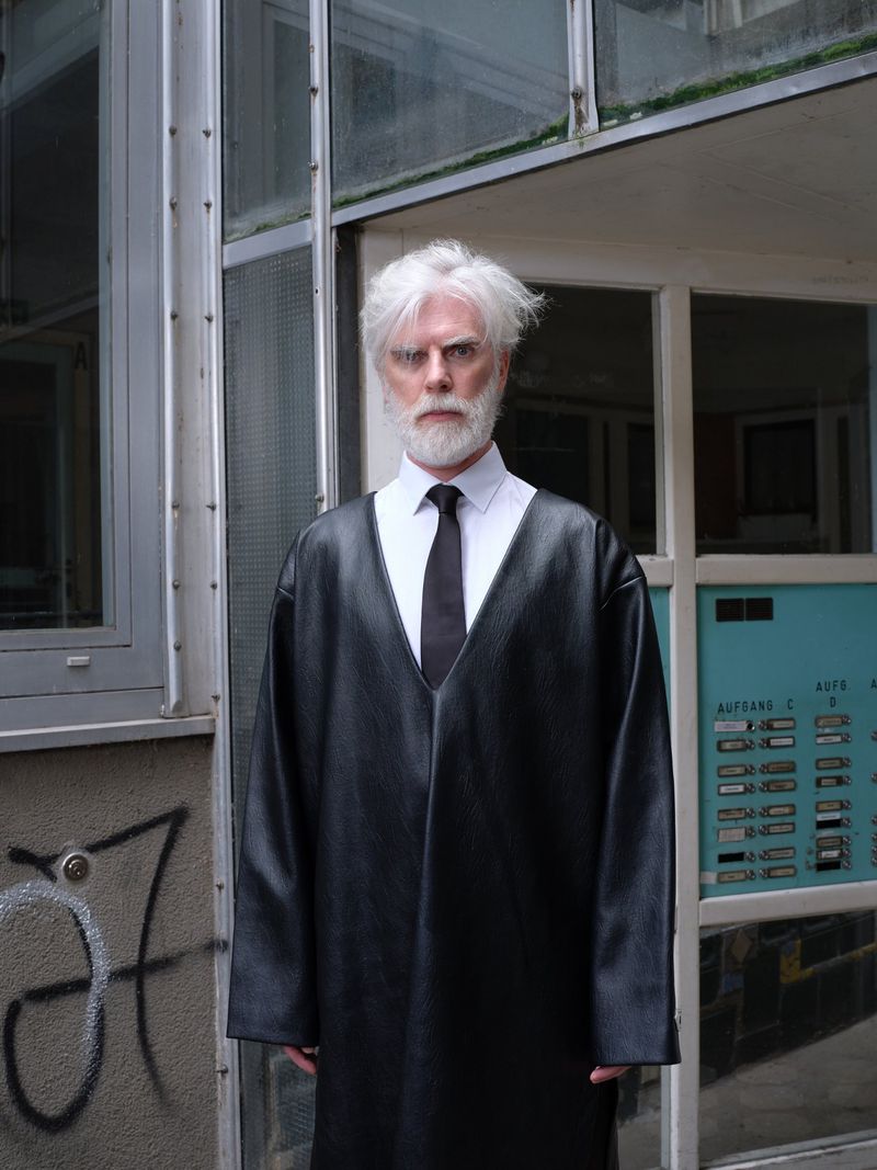 Mature adult poses outdoors wearing a black robe-like garment over a white collared shirt with a black tie against building facade.
