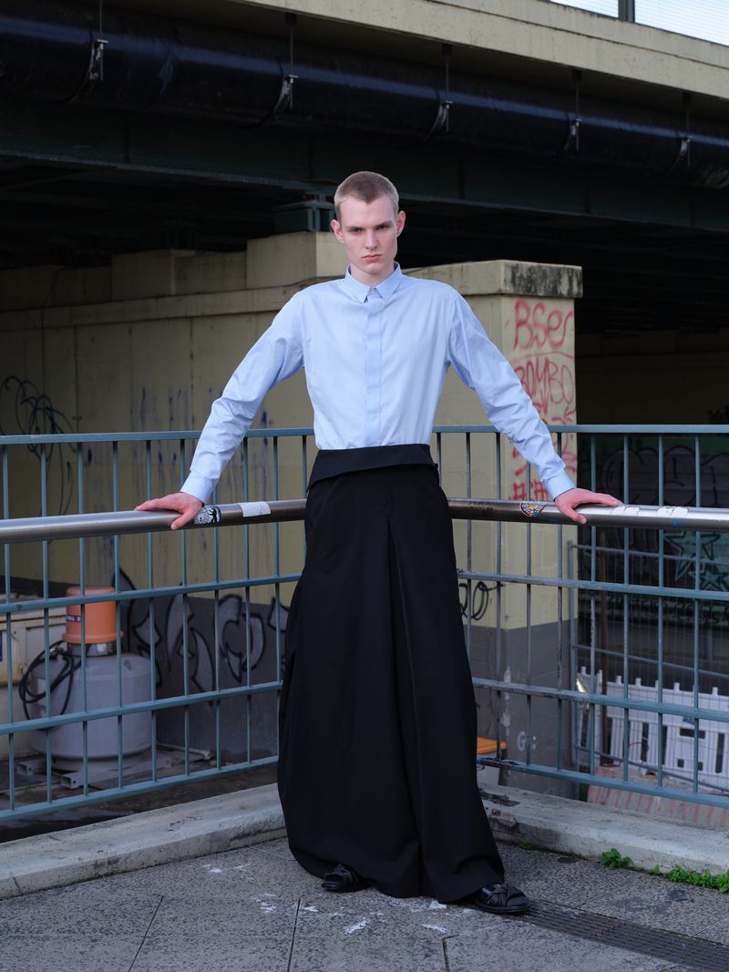 Fashionable adult male in blue shirt and black pants poses against a metal railing in an urban setting with graffiti in background.