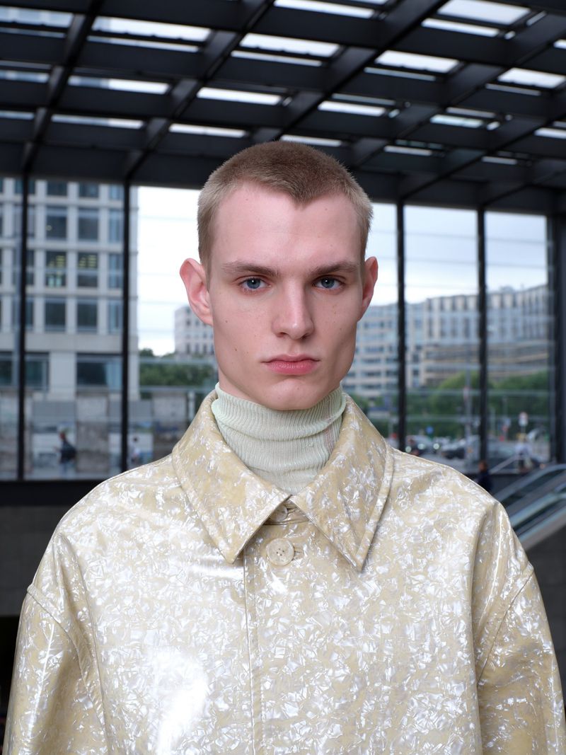 Close-up of a blonde-haired man in a glossy light-yellow coat and light-green turtleneck, set against a blurred urban background.