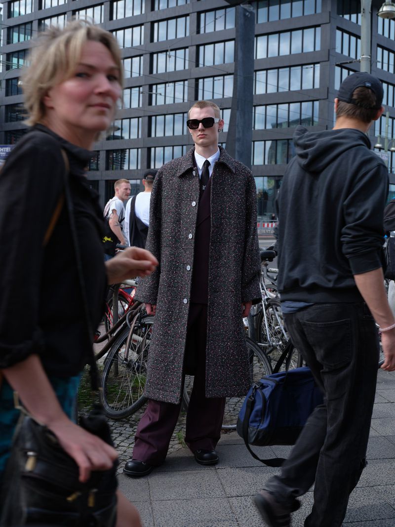A stylish adult male stands in an urban setting wearing sunglasses, a tweed coat, suit, and tie, with other people walking by.