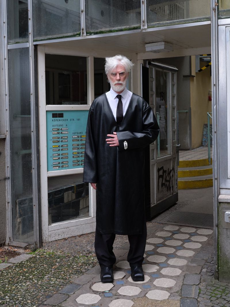 Mature adult male in a dark coat and tie standing in the doorway of a vintage building on a tiled surface.