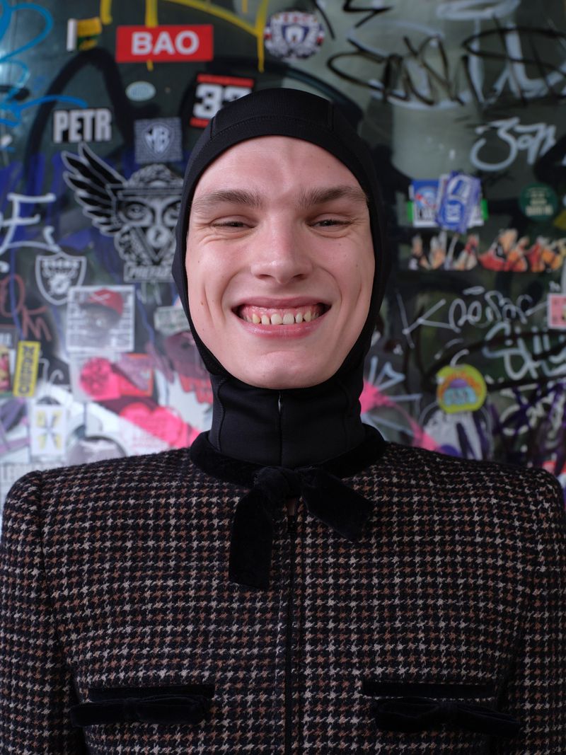 Portrait of a smiling young adult male wearing a tweed jacket with velvet trim, a black balaclava, and standing against a graffiti wall.