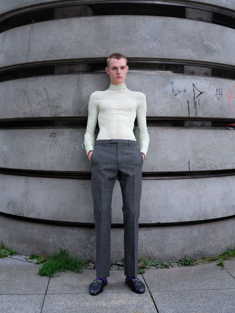 Young man in a light-green turtleneck and grey dress pants stands against a textured concrete wall in a full shot on an overcast day.