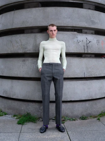 Young man in a light-green turtleneck and grey dress pants stands against a textured concrete wall in a full shot on an overcast day.