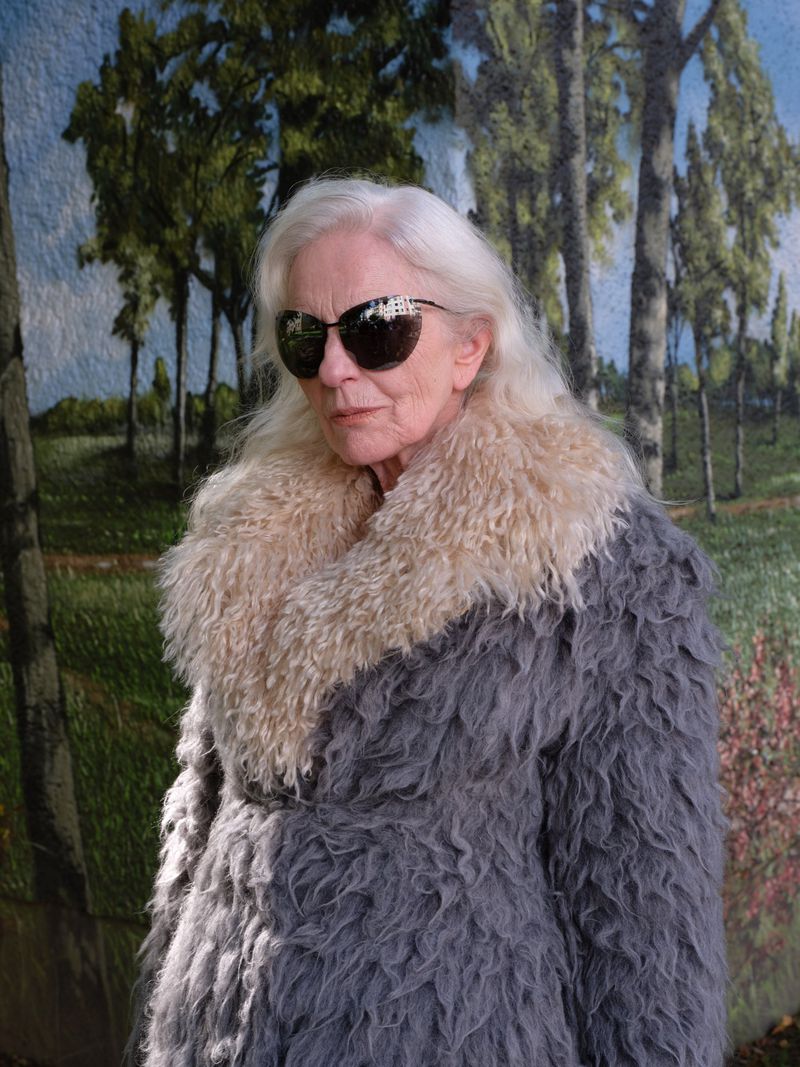 Elderly woman in fuzzy gray coat with beige fur collar and sunglasses stands in front of a mural of trees and a blue sky.