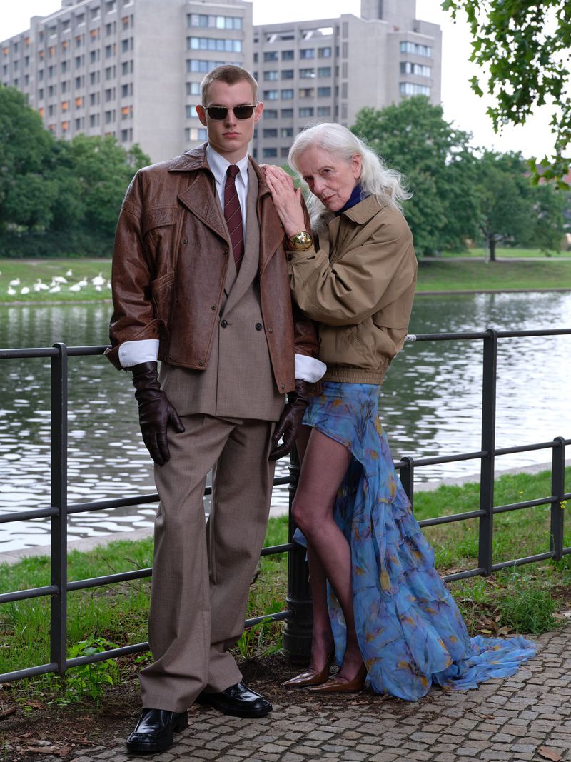 An older and younger adult posing near a pond in a fashion shot, the younger in a suit and sunglasses and the elder in a dress and jacket.