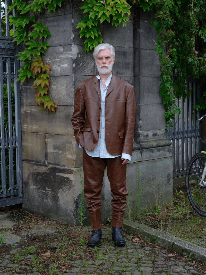 Mature man with white hair and beard, wearing a brown leather suit and boots, stands in front of a stone wall covered with ivy.