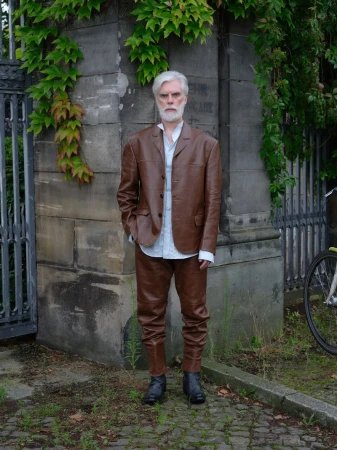 Mature man with white hair and beard, wearing a brown leather suit and boots, stands in front of a stone wall covered with ivy.