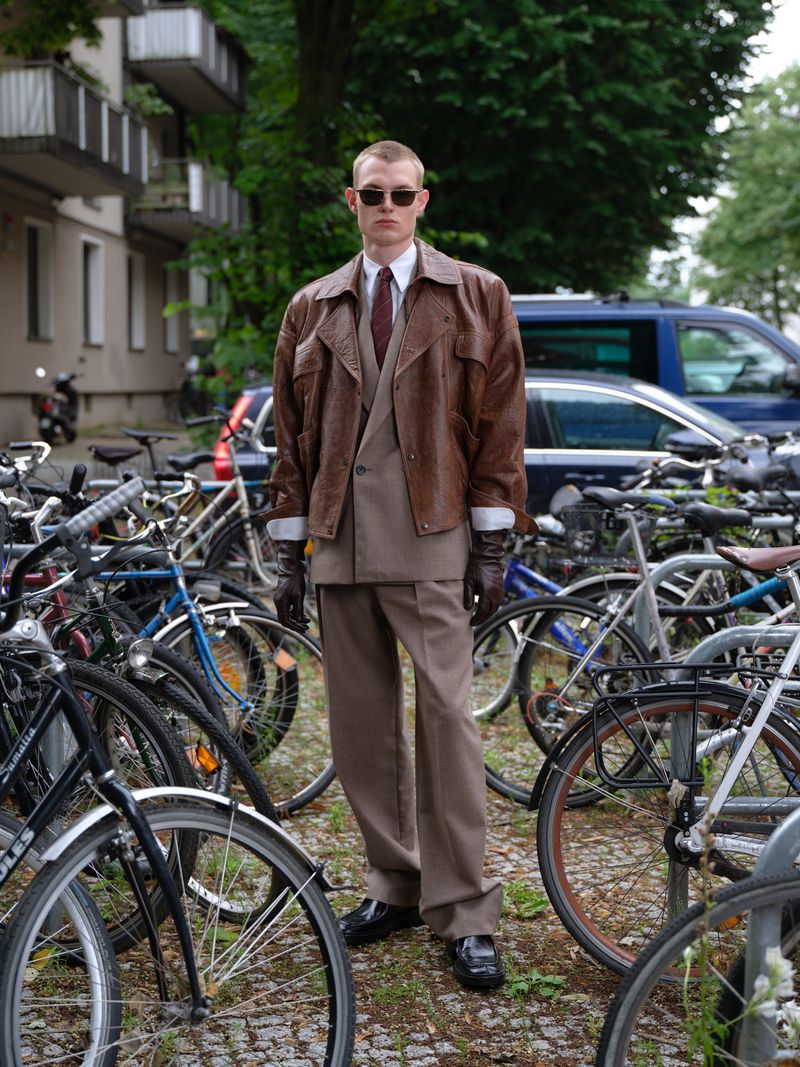 Man in suit and brown leather jacket stands among parked bicycles in an urban environment on an overcast day, wearing sunglasses.