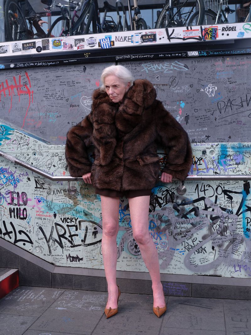 Elderly woman with short white hair wearing a voluminous brown fur coat and pointed heels posing in front of a graffiti-covered glass wall.