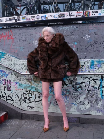 Elderly woman with short white hair wearing a voluminous brown fur coat and pointed heels posing in front of a graffiti-covered glass wall.